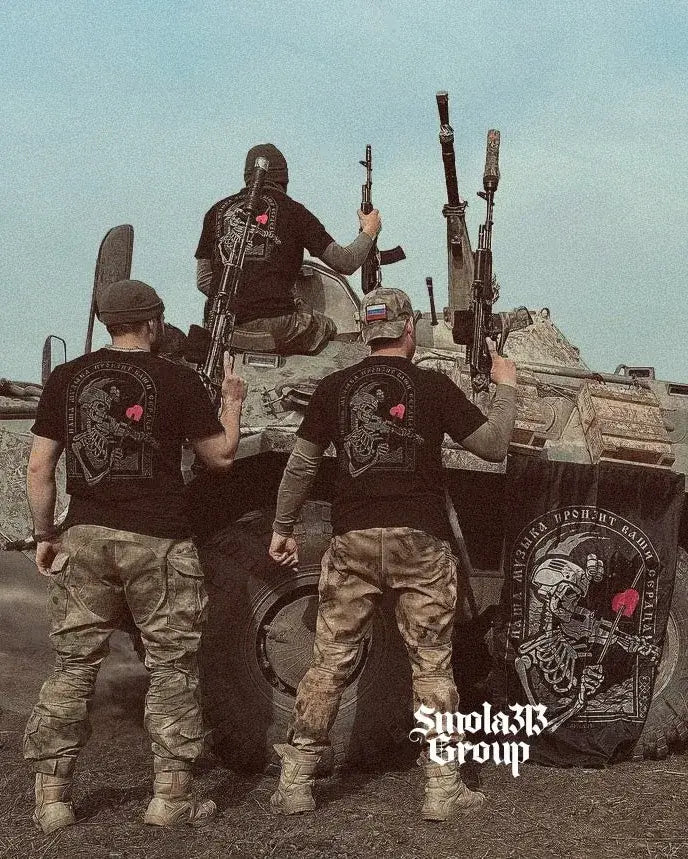 Four men in tactical gear with rifles on a military vehicle, wearing black t-shirts from Smola313 Group